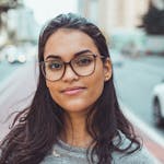 A confident woman wearing glasses smiles subtly while standing on an urban street with blurred background.