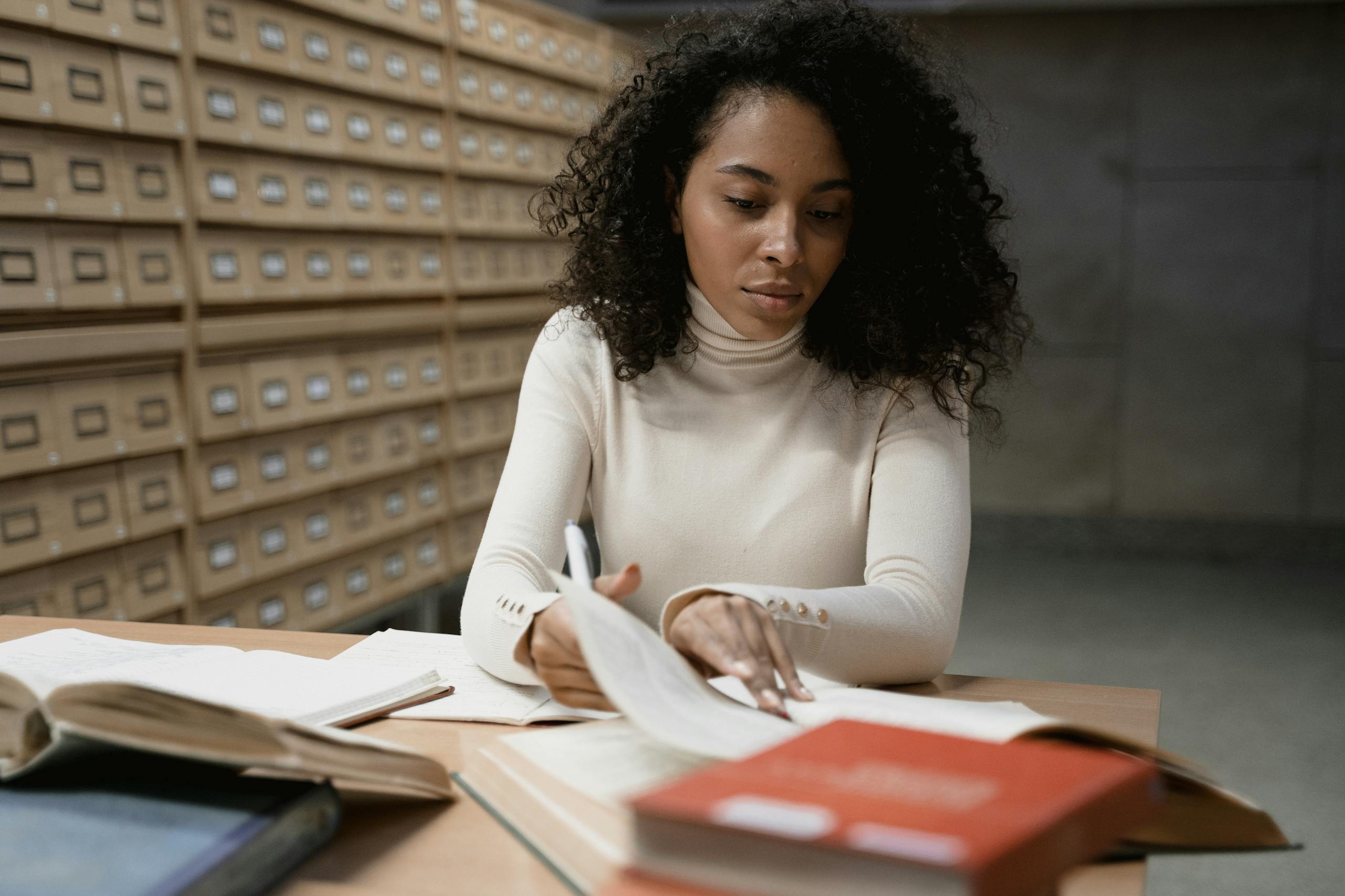 A focused young woman studying in a library, surrounded by open books and card catalog cabinets.