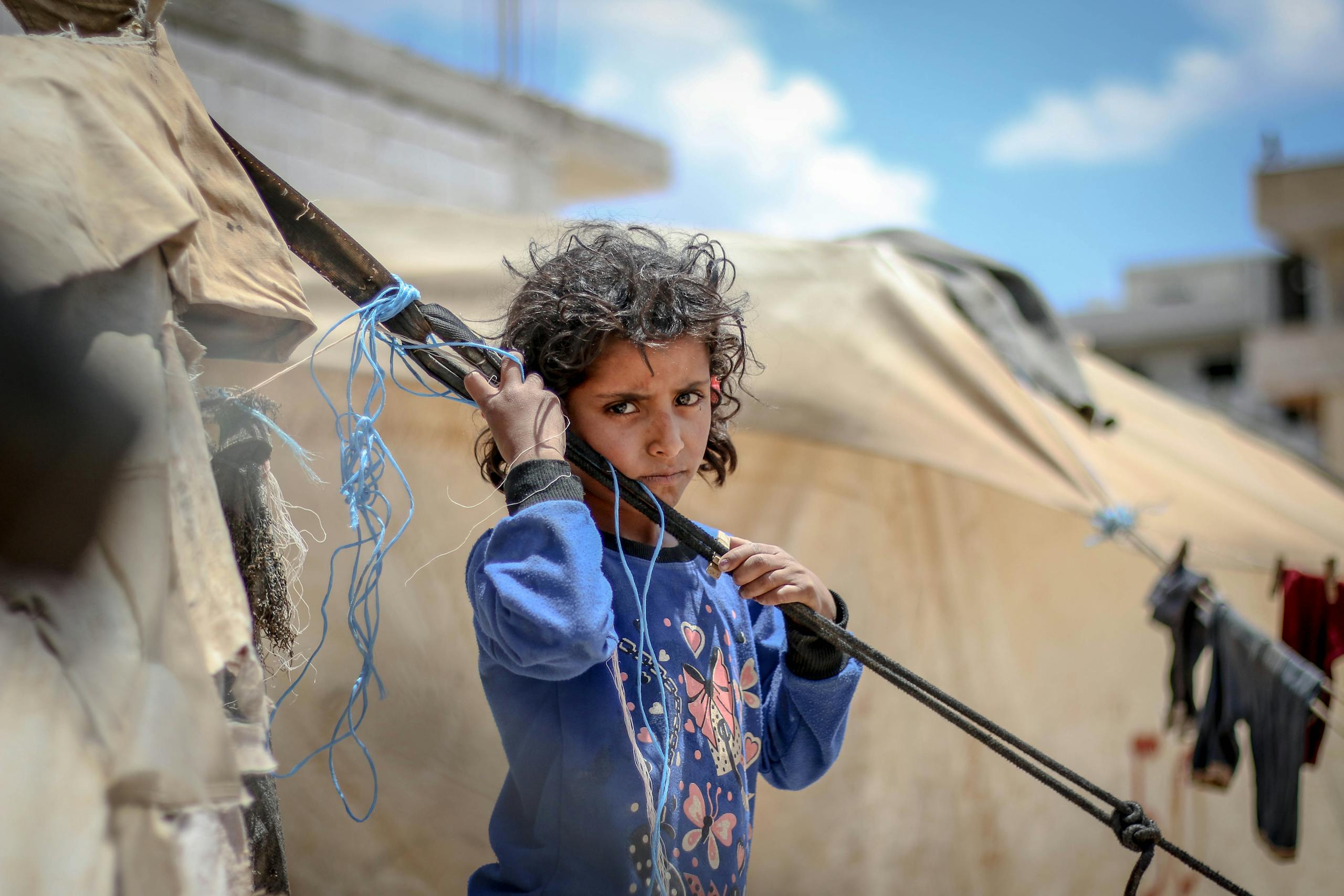 A young girl stands thoughtfully in a refugee camp, surrounded by tents and a clear sky.