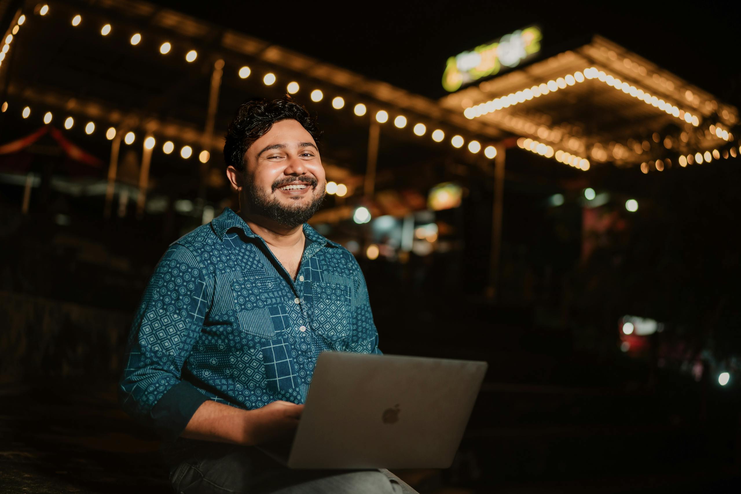 Cheerful man using laptop outdoors at night under festive lights.