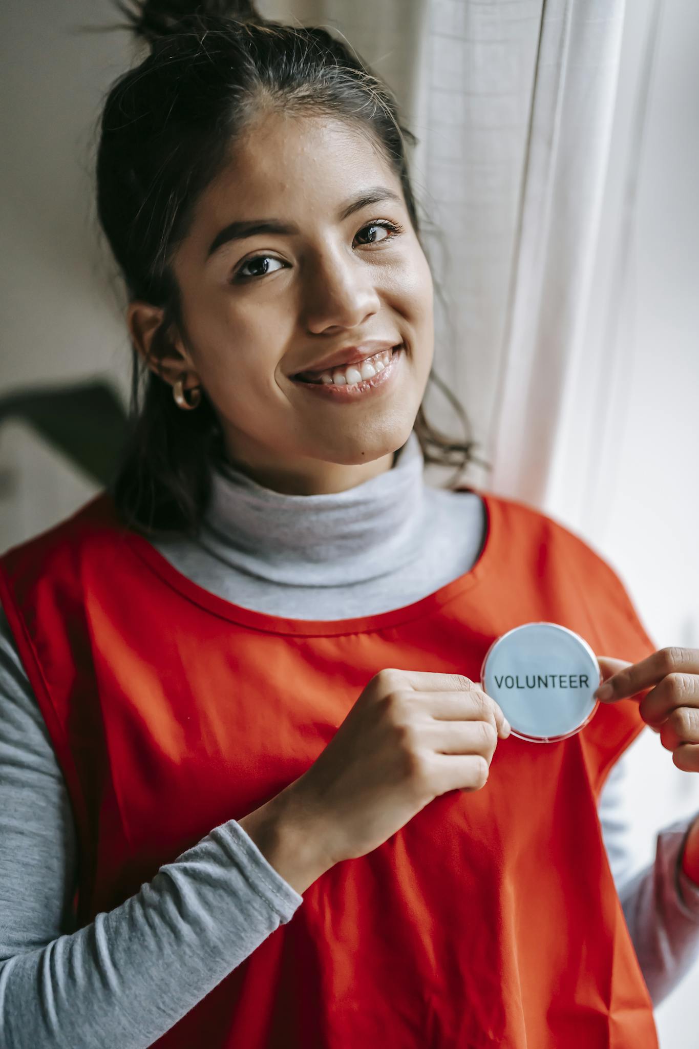 Confident female volunteer smiling while holding a volunteer badge indoors.