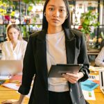 Confident professional woman holding a tablet in a vibrant modern office with colleagues working in the background.