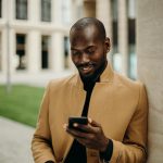 Relaxed man in coat smiles while using smartphone and holding coffee outside.