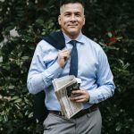 Smiling man in formal wear posing with a newspaper in an outdoor setting with greenery.