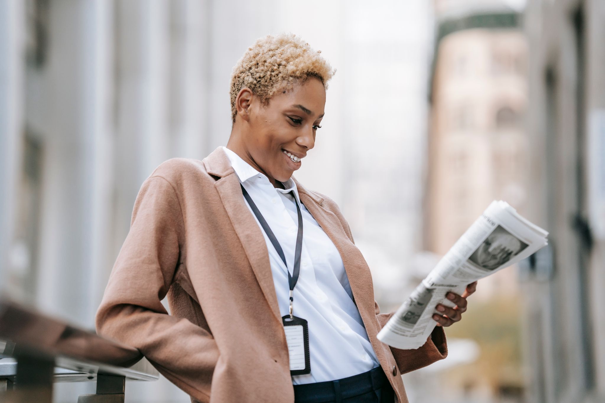 Delighted young African American female entrepreneur in stylish brown coat with badge leaning on railing while reading newspaper