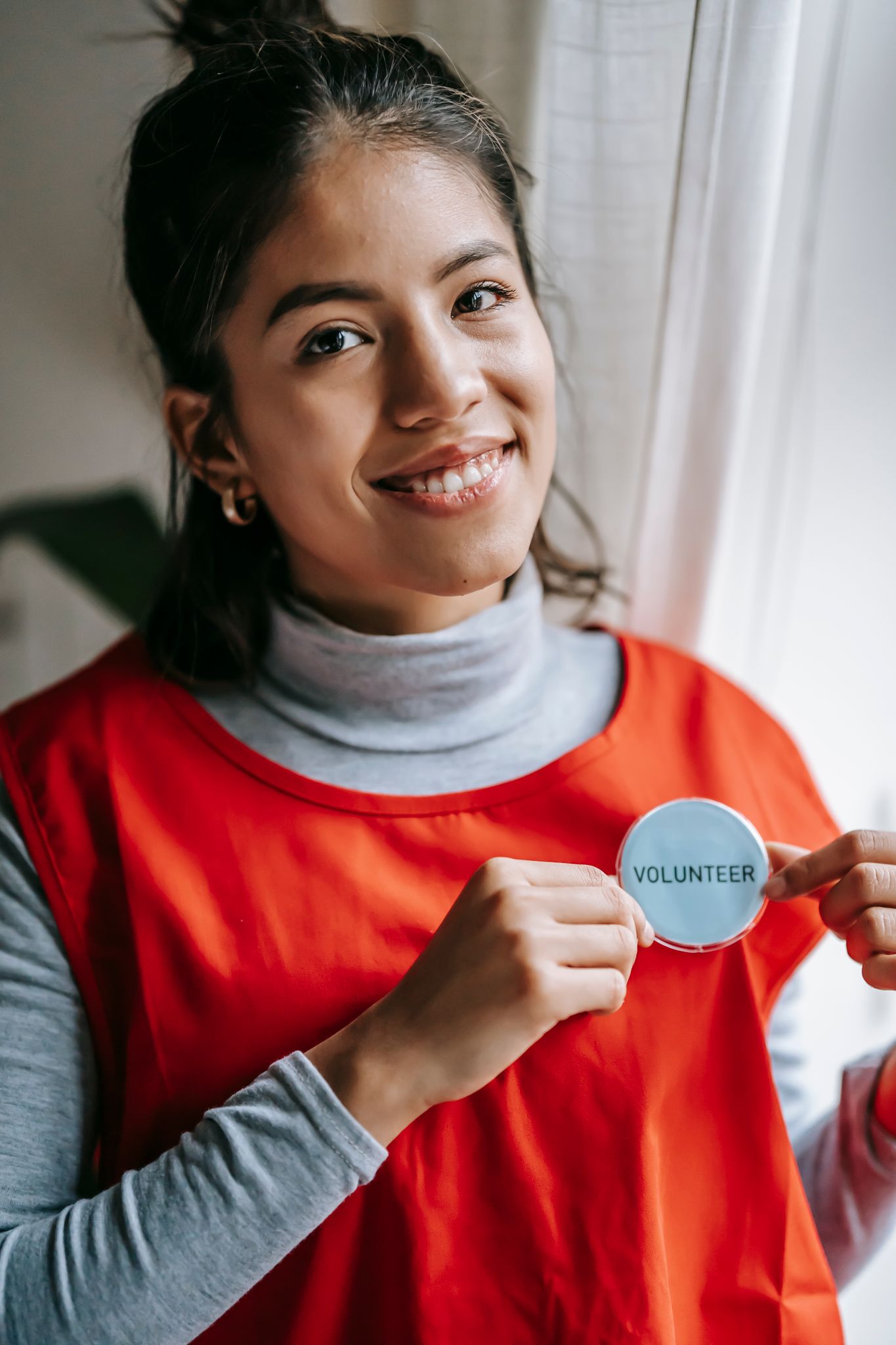Happy ethnic female volunteer in casual outfit and red uniform looking at camera and smiling while demonstrating badge with inscription in light room